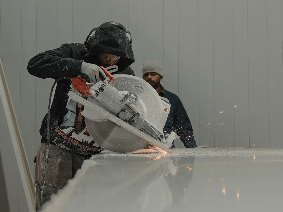 WFE worker in branded black hoodie cutting a large insulated panel with a circular saw at the 35 Silton site, sparks flying from the blade, a second crew member watching in the background.