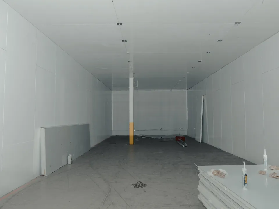 Near-finished walk-in cold storage interior at 35 Silton: long white insulated panel envelope floor to ceiling, a yellow-wrapped structural support column running through the middle of the room, sealant tubes staged on finished panels at right.