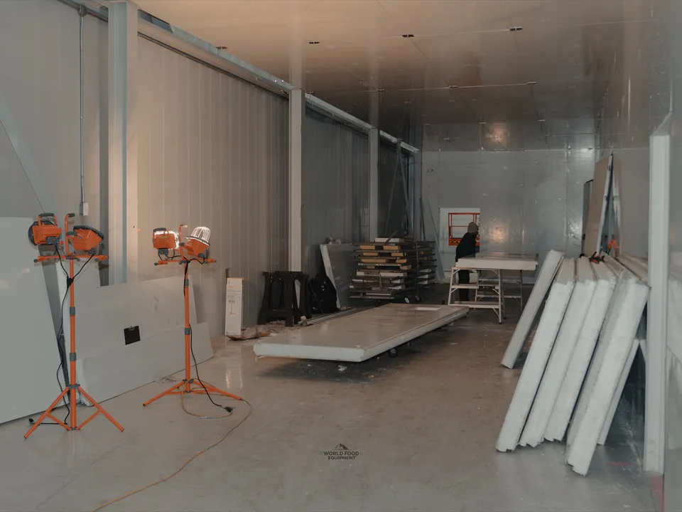 Active build zone inside the 35 Silton cold storage envelope: three orange Milwaukee work-light tripods staged on the floor, stacked insulated panels along the wall, a WFE worker visible mid-distance at a sawhorse, white insulated panel walls overhead.