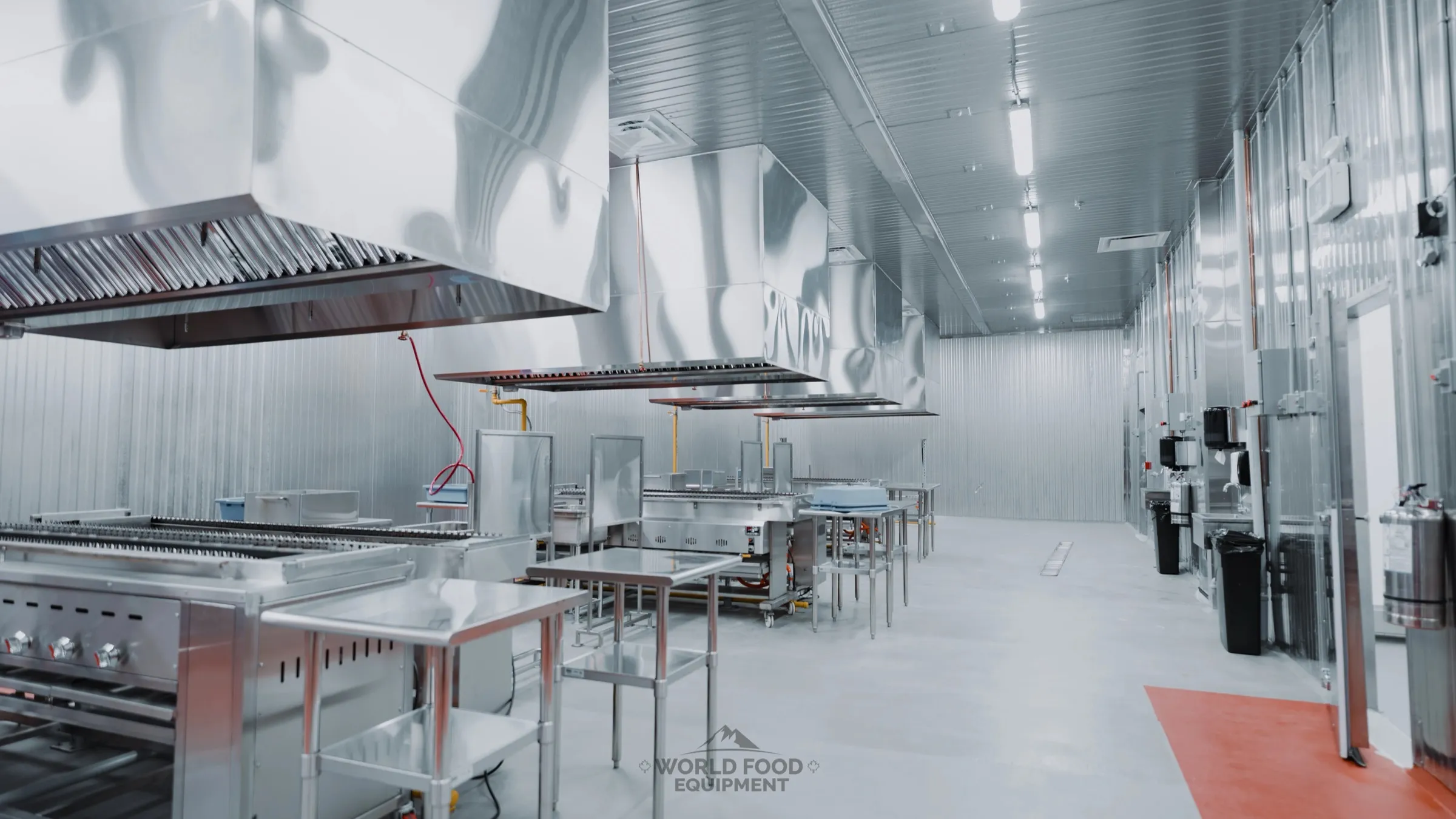 Wide commercial restaurant kitchen interior with a bank of stainless steel chef hoods over the prep line, stainless prep tables, vertical stainless storage panels on the side walls, polished concrete floor with a red drain channel under LED ceiling strips, Al Shamas restaurant kitchen by World Food Equipment in Ontario
