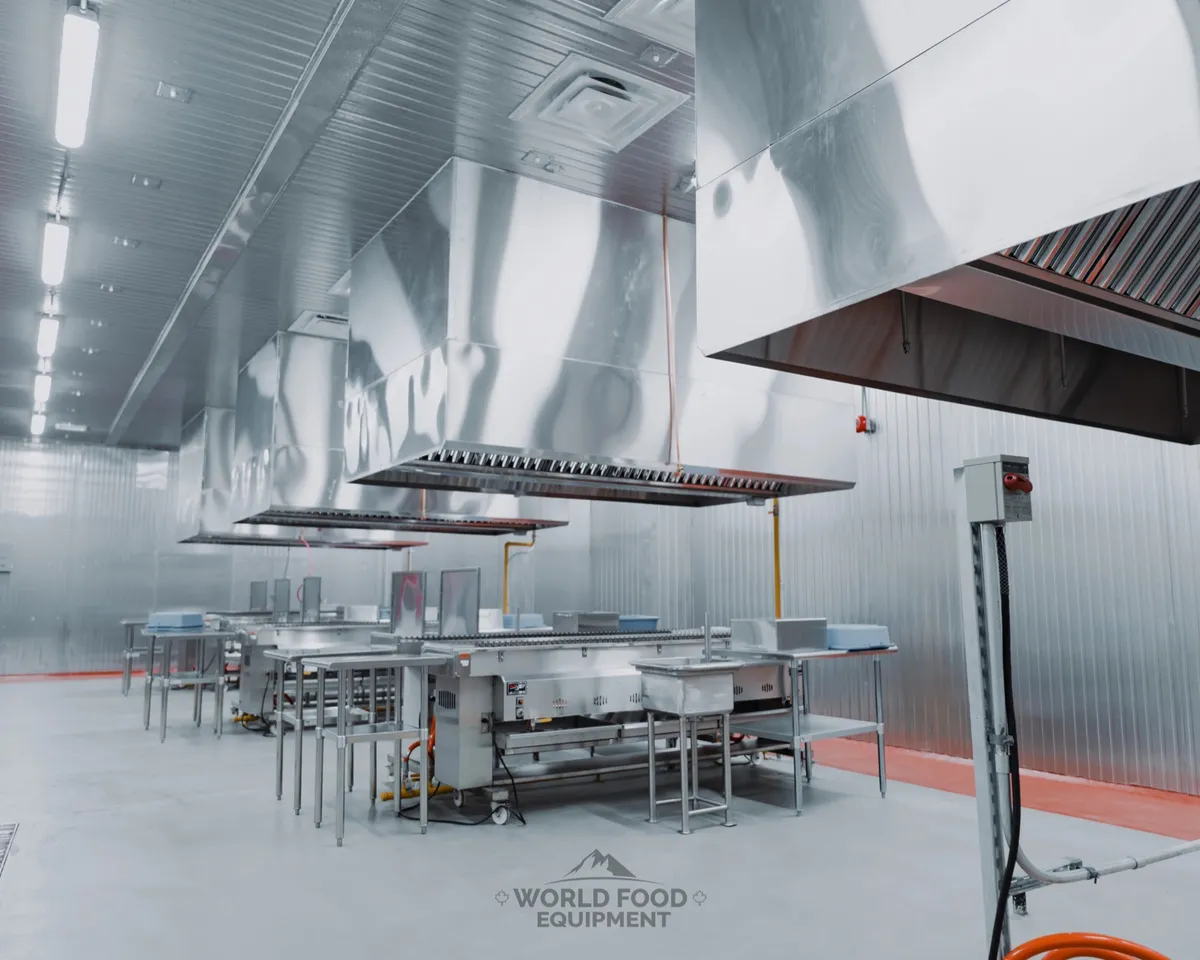 Finished commercial restaurant kitchen with full stainless-steel wall cladding, suspended stainless exhaust hoods over a central cooking line, stainless prep tables and a recessed floor drainage channel, polished concrete floor, Al Shamas commercial kitchen install by World Food Equipment