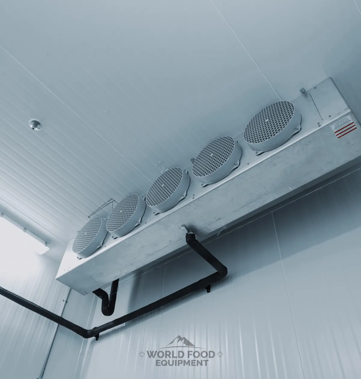 Five-fan ceiling-mounted evaporator in the Al Shamas Food Products cold room, galvanized housing against white insulated ceiling panels, insulated drain line run along the wall, vapor-tight light fixture beside it.