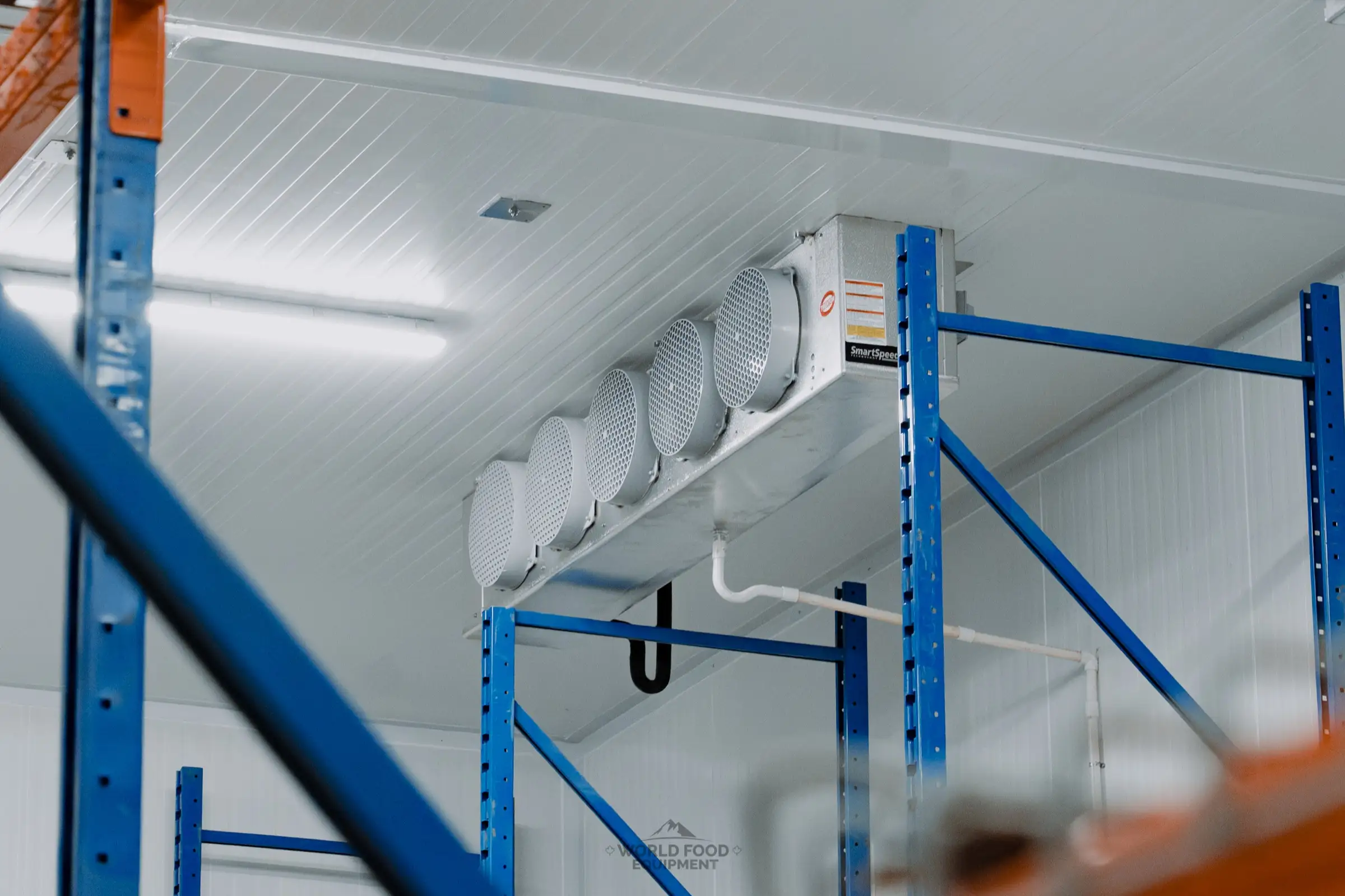 Five-fan ceiling evaporator mounted above blue pallet racking uprights inside the finished Atlas Foods cold storage warehouse in Toronto, white insulated ceiling panels and condensate drain line, World Food Equipment install