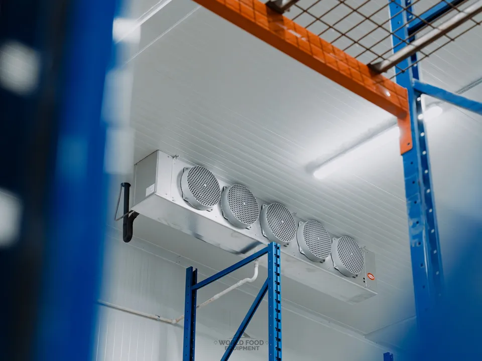First five-fan ceiling-mounted evaporator hung against the Atlas Foods cooler ceiling with blue pallet racking framing the foreground and a refrigeration line running down to the condenser below.