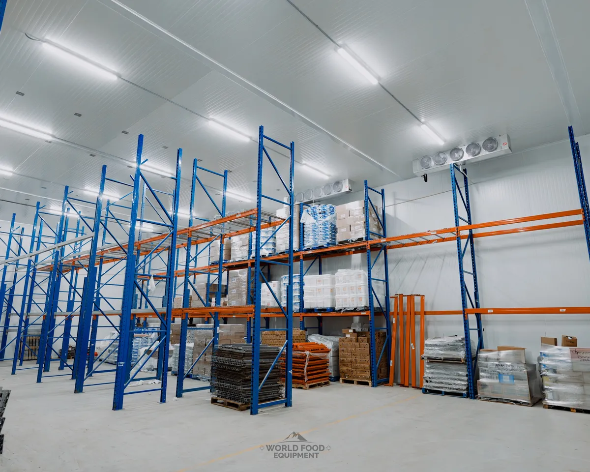 Stocked commercial cold storage room with blue and orange industrial pallet racking loaded with product, a ceiling-mounted evaporator on the back wall, white insulated panel walls, polished concrete floor, Atlas Foods install, Ontario commercial cold storage by World Food Equipment
