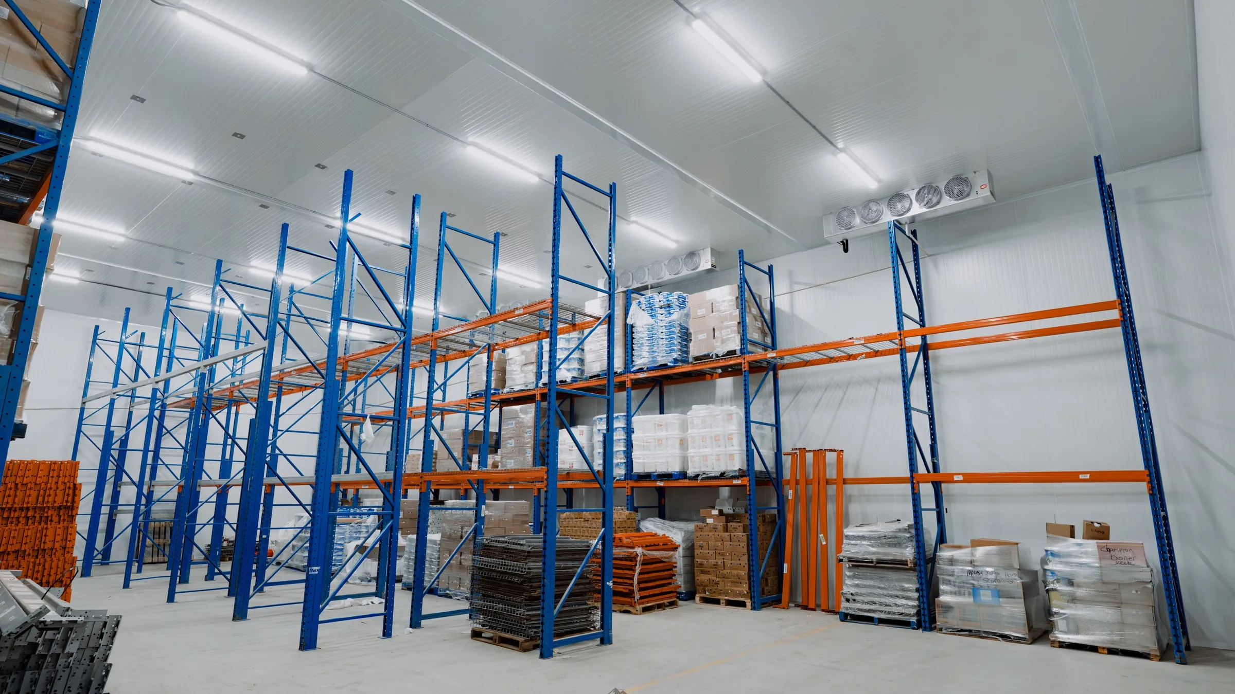 Finished commercial cold storage warehouse in Ontario with tall blue and orange pallet racking, two ceiling-mounted multi-fan evaporators, white insulated panel walls and ceiling, palletized product staged on the floor, Atlas install by World Food Equipment