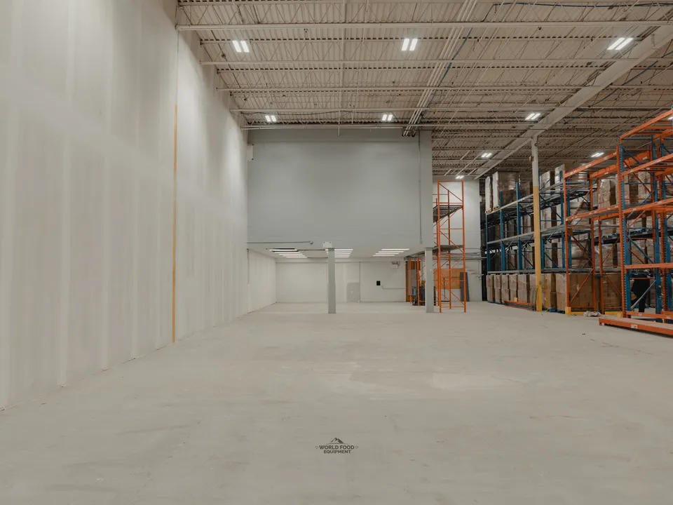 Atlas Foods cold storage envelope nearly complete in week two, central structural support column anchored in the middle of the room, doorway openings framed at the back wall, orange pallet racking running along the right side.