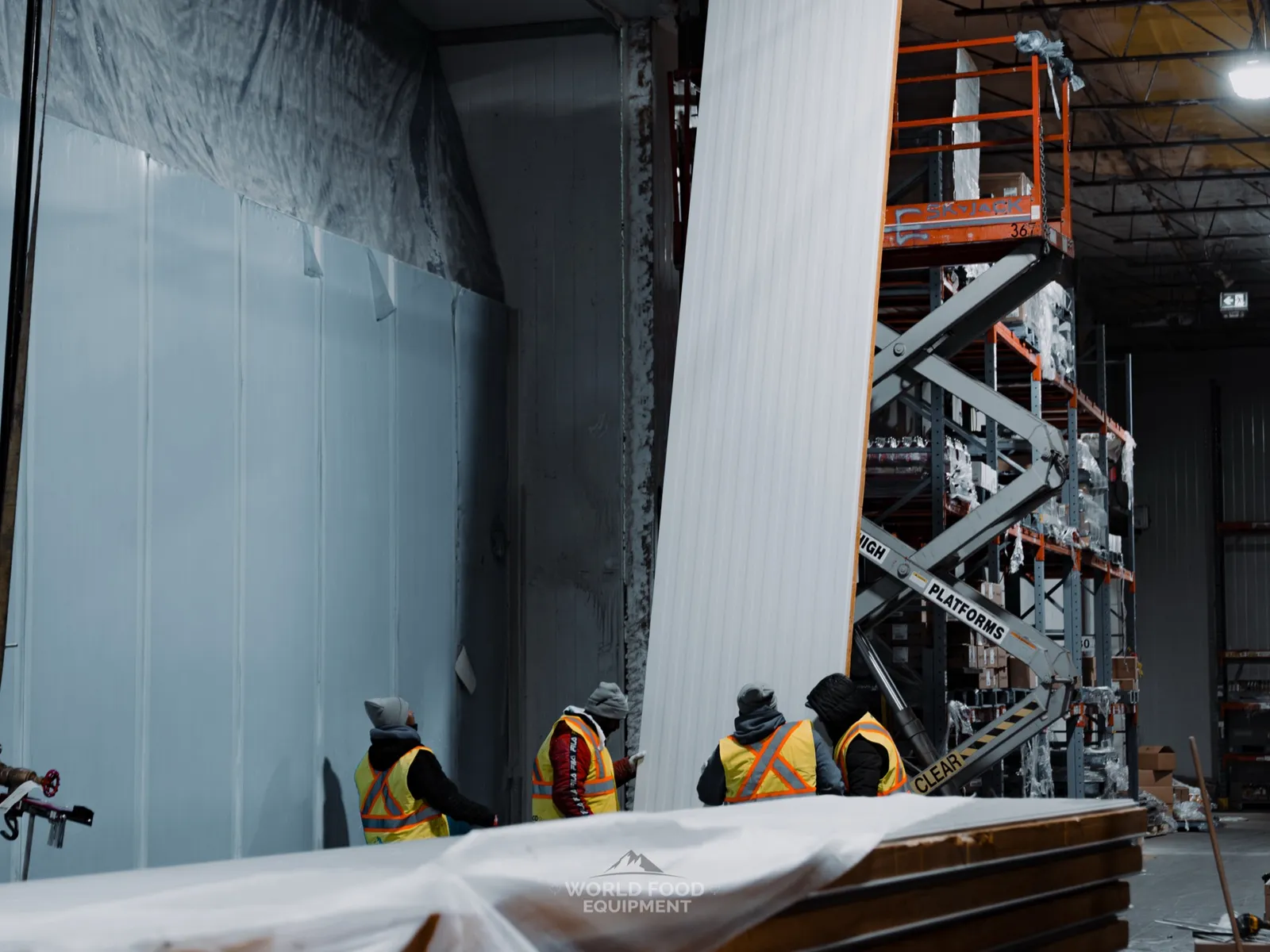 Four WFE crew at CJR Grocers positioning a tall white insulated freezer-wall panel mid-lift, scissor lift staged behind them, vapor-barrier wall visible to the left where the failed 140 ft section came out, grocery racking visible to the right.