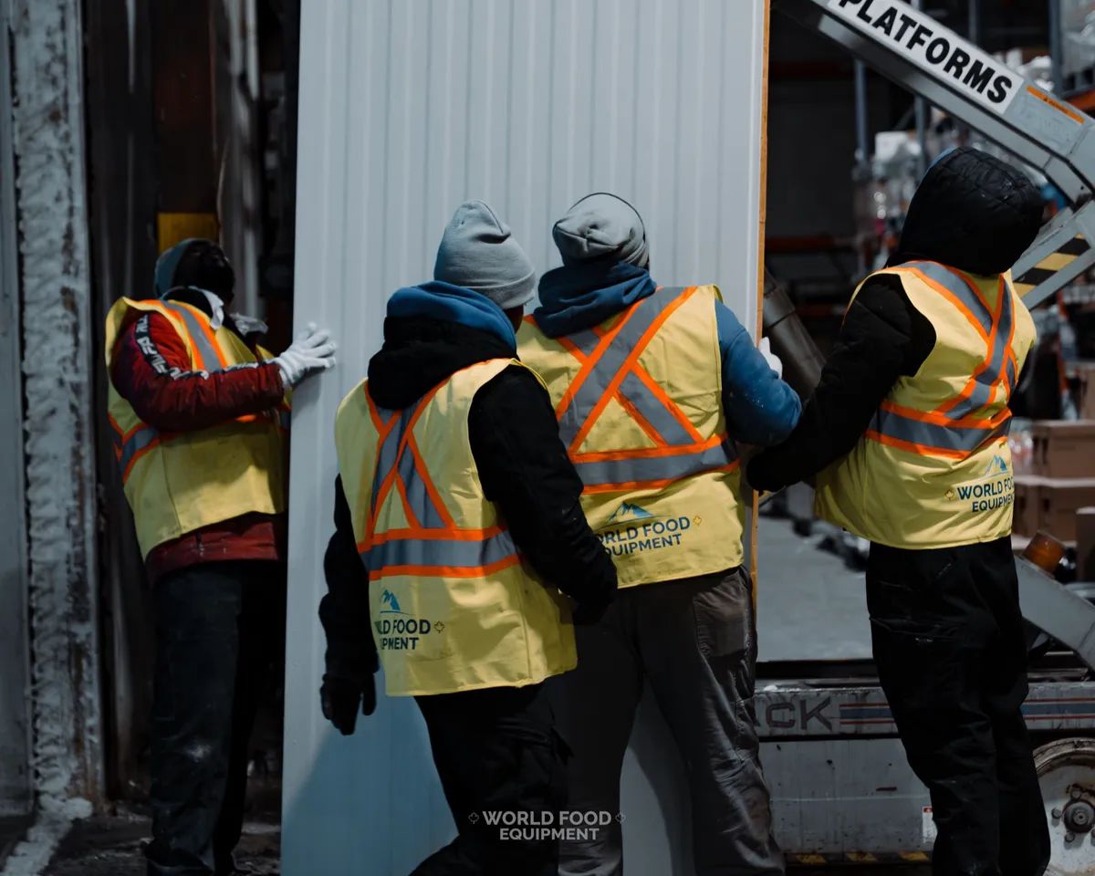 Four World Food Equipment crew members in branded hi-vis safety vests carrying a large white insulated wall panel into position during a commercial cold storage install, pallet racking in the background, Ontario installation by World Food Equipment
