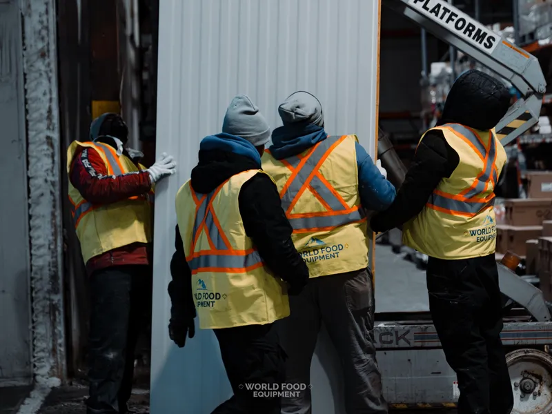 Four WFE crew in hi-vis vests pushing a tall white insulated freezer-wall panel into place at CJR Grocers, panel against the rebuilt base, grocery racking visible in the background.