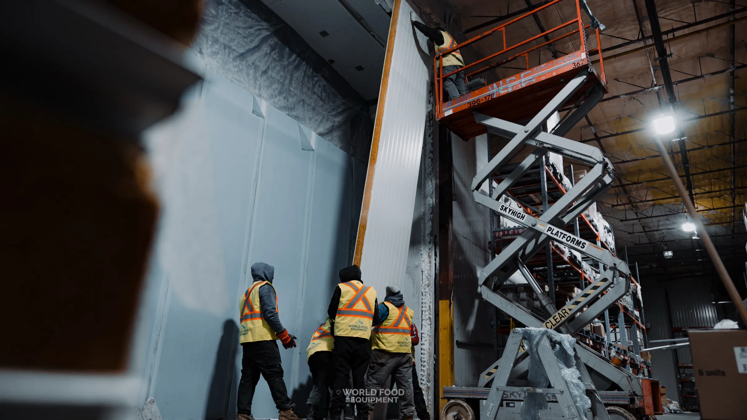 Three World Food Equipment installers in branded high-visibility safety vests viewed from behind, pushing a tall narrow insulated cold-storage panel into place, scissor lift staged on the right, stocked grocery product visible on warehouse pallet racking in the background, CJR Grocers install, Ontario commercial walk-in installation by World Food Equipment