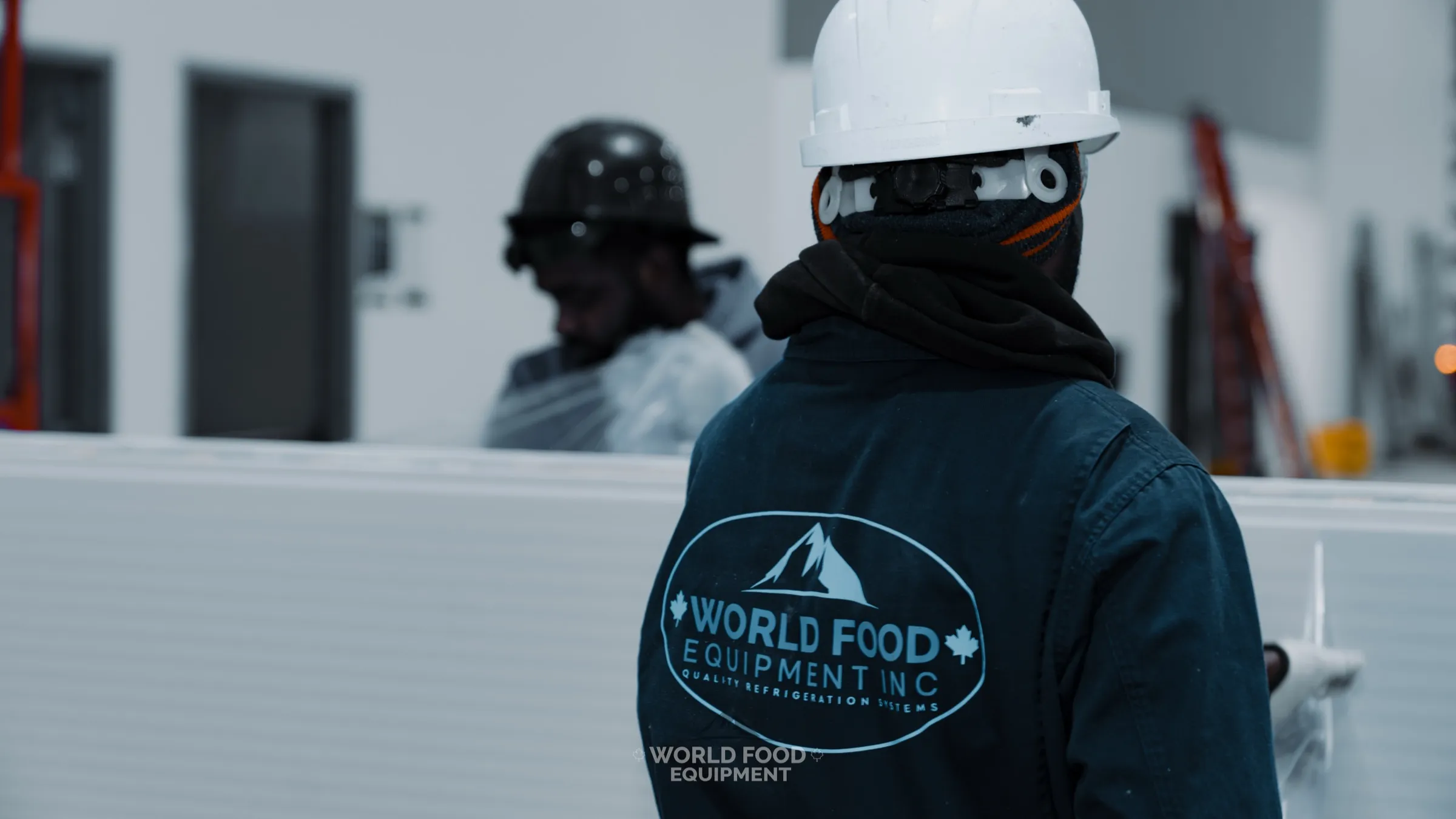 World Food Equipment Inc installer in branded dark navy WFE coveralls with the oval mountain logo and Quality Refrigeration Systems wordmark across the back, white hard hat, second WFE crew member in a black helmet behind, positioning a white insulated panel on a commercial cold storage build, Chocolate Factory install, Ontario, World Food Equipment