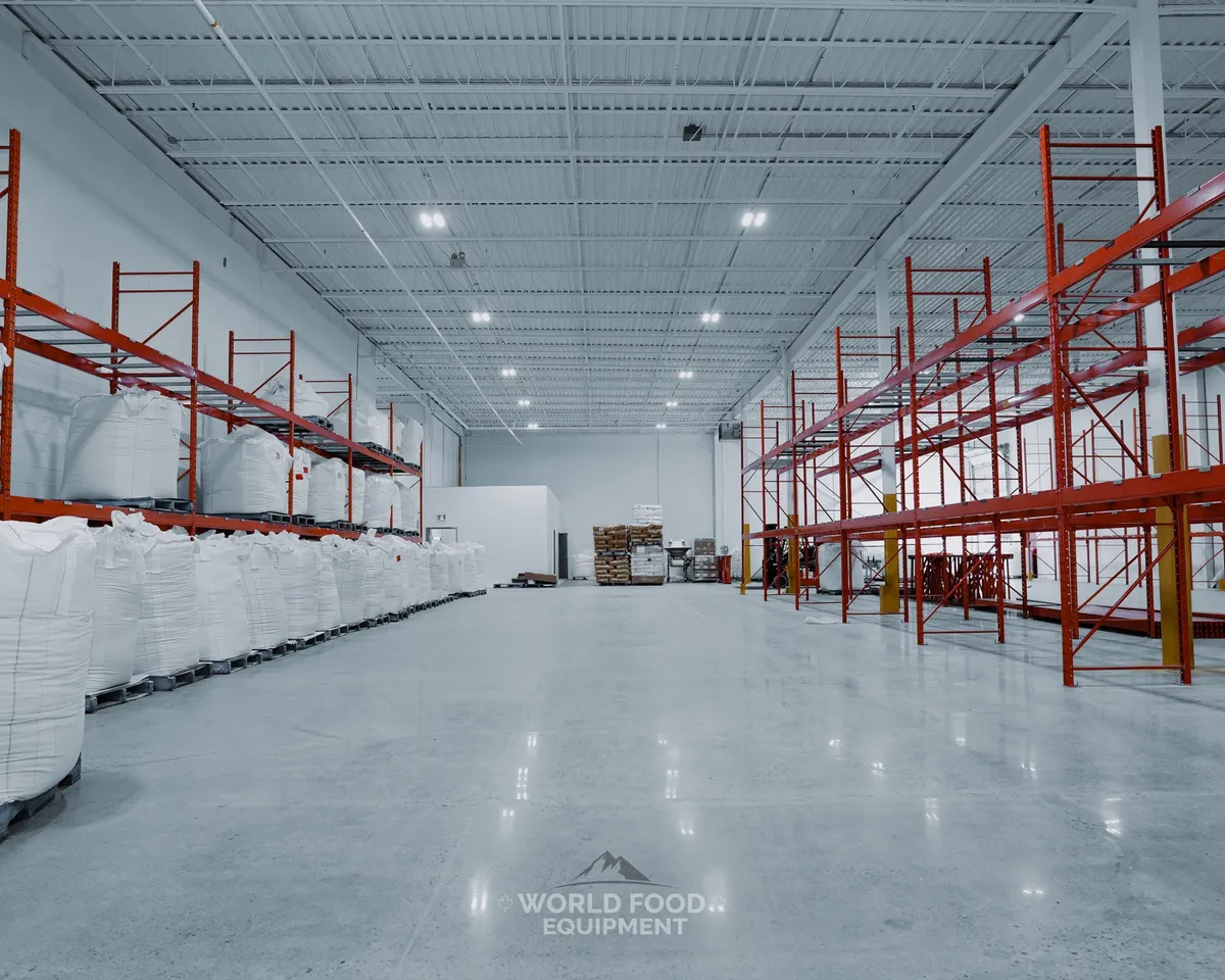 Wide industrial distribution warehouse with tall orange pallet racking down the right side, white super-sacks of bulk material stacked on the left, a forklift on the polished concrete floor, a finished white insulated cold-storage envelope in the back center, Ontario commercial cold storage install by World Food Equipment