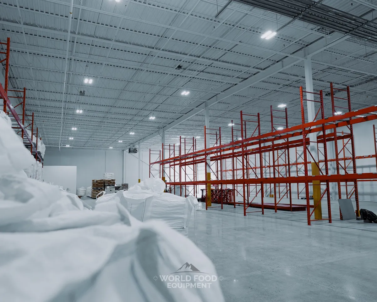 Warehouse-scale cold storage interior with tall orange-and-blue industrial pallet racking down a wide aisle, stacked bulk product, scissor lift, bright overhead lighting under steel trusses, Chocolate Factory install, Toronto, World Food Equipment