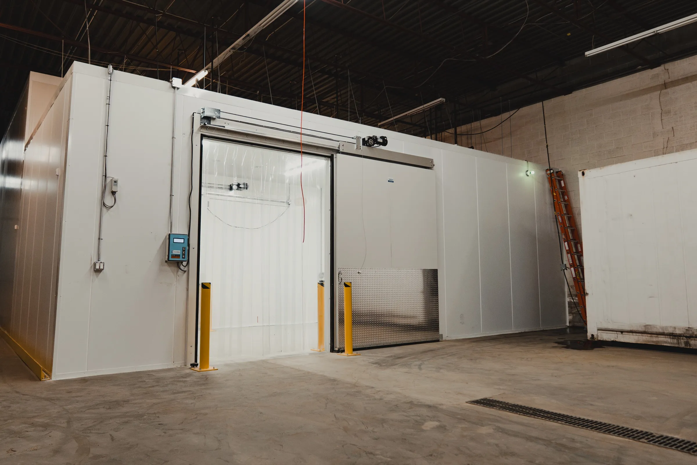Exterior of the finished SKOR Crestlawn walk-in freezer envelope built by World Food Equipment in Ontario. Large white insulated panel envelope with a wide automatic sliding insulated freezer door fronted by twin yellow steel bollards, controls box and digital temperature panel on the left wall, polished concrete warehouse floor
