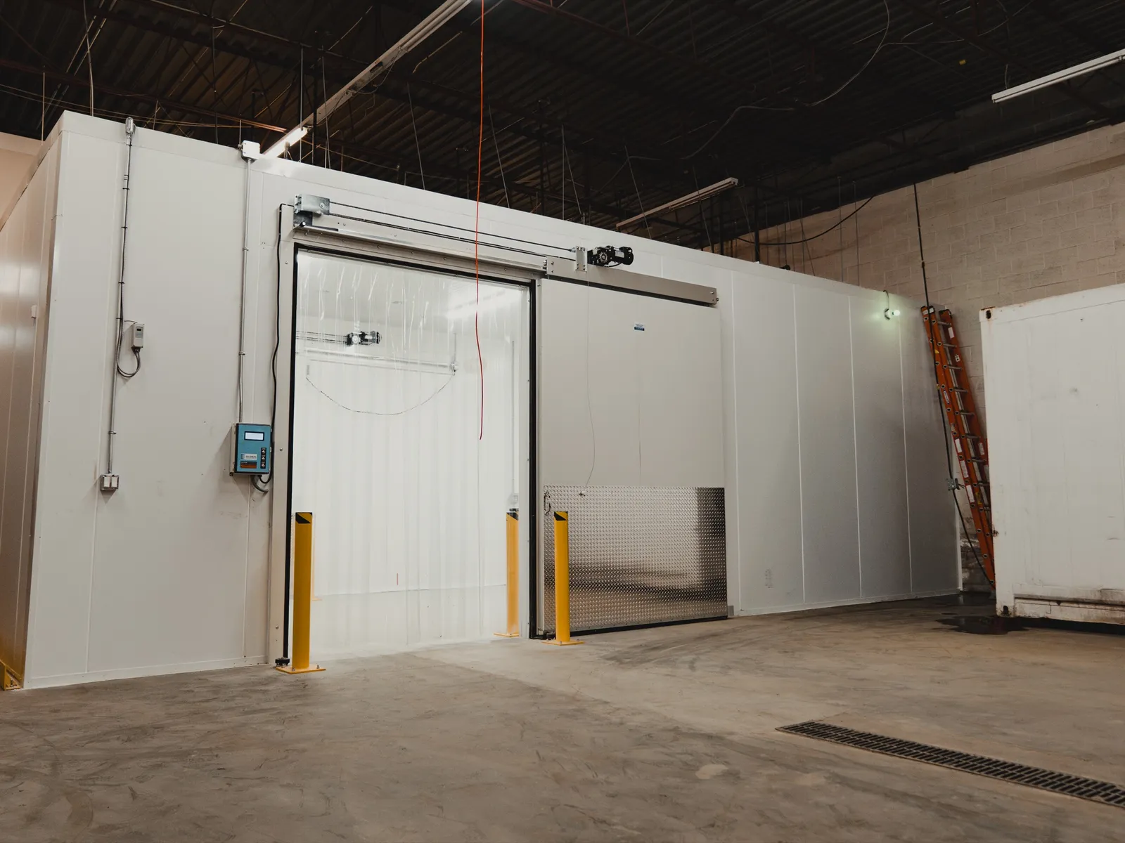 Walk-in freezer envelope at SKOR Crestlawn dropped into the larger warehouse facility: white-panel cold storage box with the sliding insulated door open, yellow steel bollards flanking the entry, darker industrial warehouse visible around the envelope, polished concrete floor with center drain.