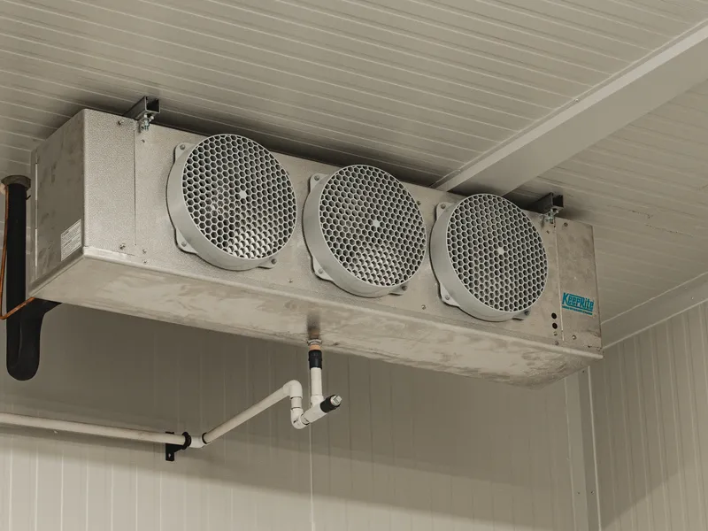 KeepRite three-fan ceiling-mounted evaporator close-up inside the finished SKOR Crestlawn walk-in freezer, KeepRite blue logo visible on the unit casing, white insulated panel ceiling above, refrigeration drain line wrapped in black insulation tape running down to a U-bend below the unit.