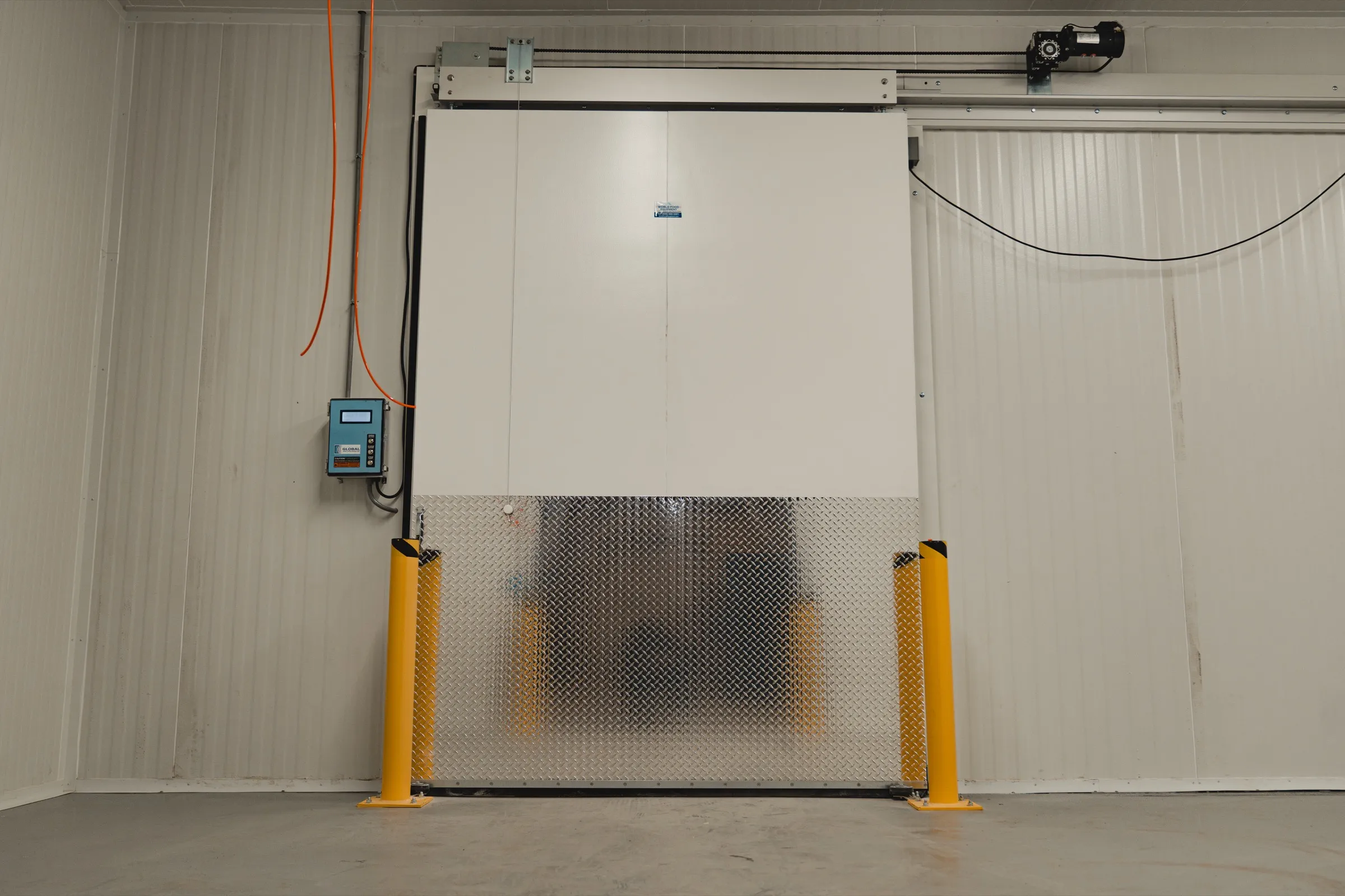 Motorized sliding insulated freezer door at the SKOR Crestlawn install in Ontario, door operator motor on the top rail, wall-mounted door controller in conduit, diamond-plate kickplate and yellow bollards, World Food Equipment