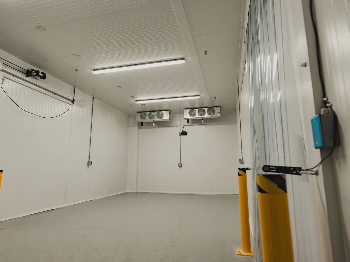 SKOR Crestlawn 3,000 sq ft walk-in freezer at handoff, dual ceiling-mounted evaporators visible against white insulated panel ceiling, yellow steel bollards staged at the doorway, refrigeration commissioned and ready for stocked production.
