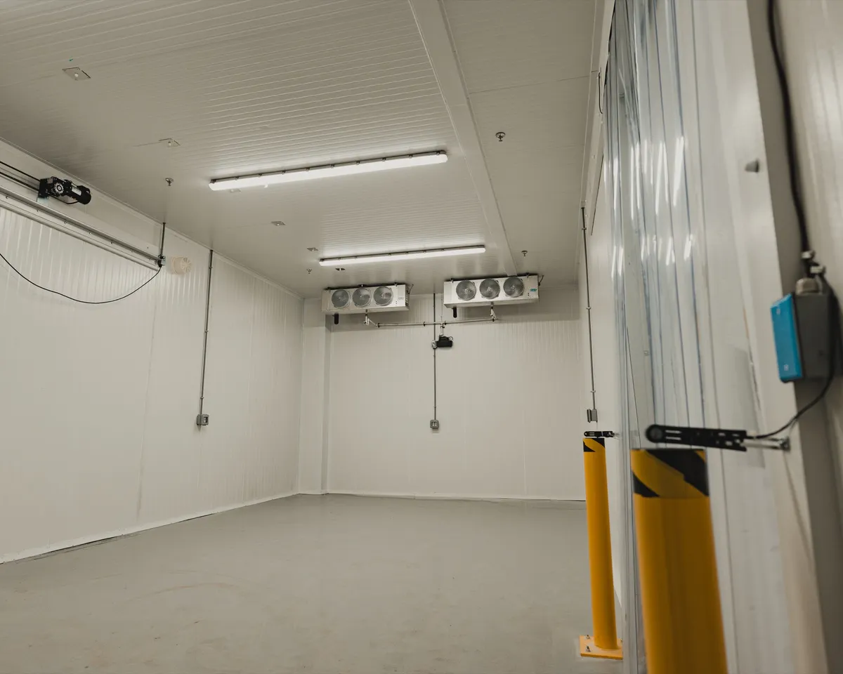 Finished commercial cold storage room with white insulated panel walls, two ceiling-mounted three-fan evaporators on the back wall, a sliding insulated door on the right with yellow steel bollards, polished concrete floor, Ontario commercial cold storage by World Food Equipment