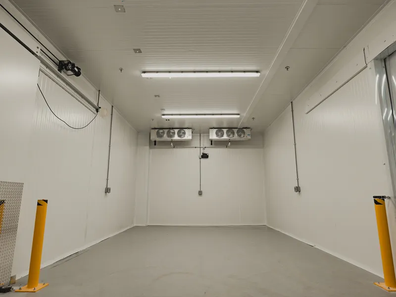 Empty walk-in freezer interior at SKOR Crestlawn looking into the room from the doorway, dual ceiling-mounted evaporators on the far wall, yellow steel bollards flanking the entry on both sides, white insulated panel walls and ceiling, polished concrete floor.