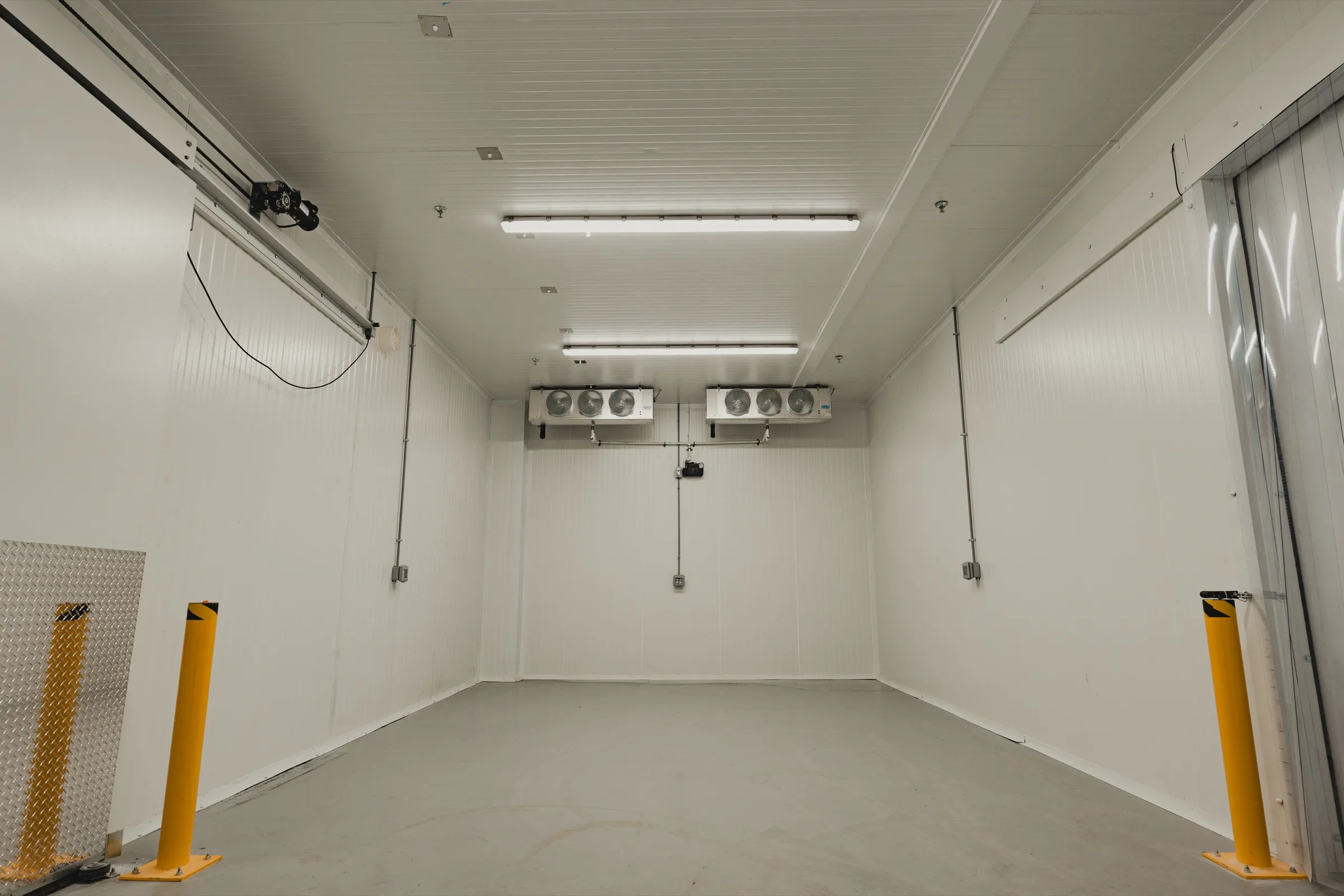 Interior of a finished walk-in cooler with two ceiling-mounted evaporators, white insulated panel walls, polished concrete floor, and yellow safety bollards on either side of the entry, Crestlawn install, Ontario, World Food Equipment