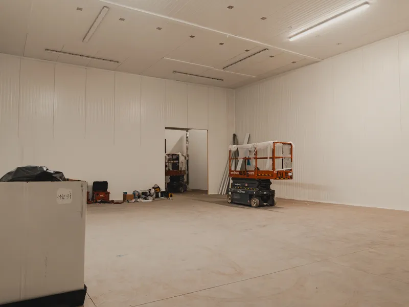 Nov 7 build phase at SKOR Crestlawn: walk-in freezer envelope already built with white insulated panel walls and ceiling, equipment and materials staged on the floor for the next phase of work, scissor lift visible to the right, second smaller room visible through an interior doorway.