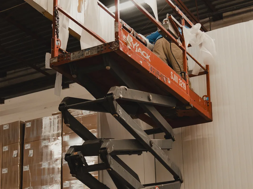 WFE worker on a fully-extended SkyJack scissor lift at SKOR Crestlawn working overhead inside the freezer envelope during the Nov 7 build phase, white insulated panel walls visible below, school food supplier facility.