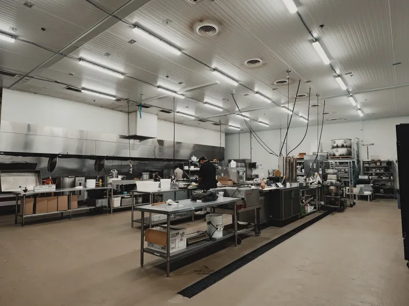 Commercial prep kitchen at Food Junction Toronto: stainless steel prep tables down the center, overhead stainless exhaust hoods running the length of the line, worker prepping food at the back, fluorescent ceiling lights, full kitchen operational.