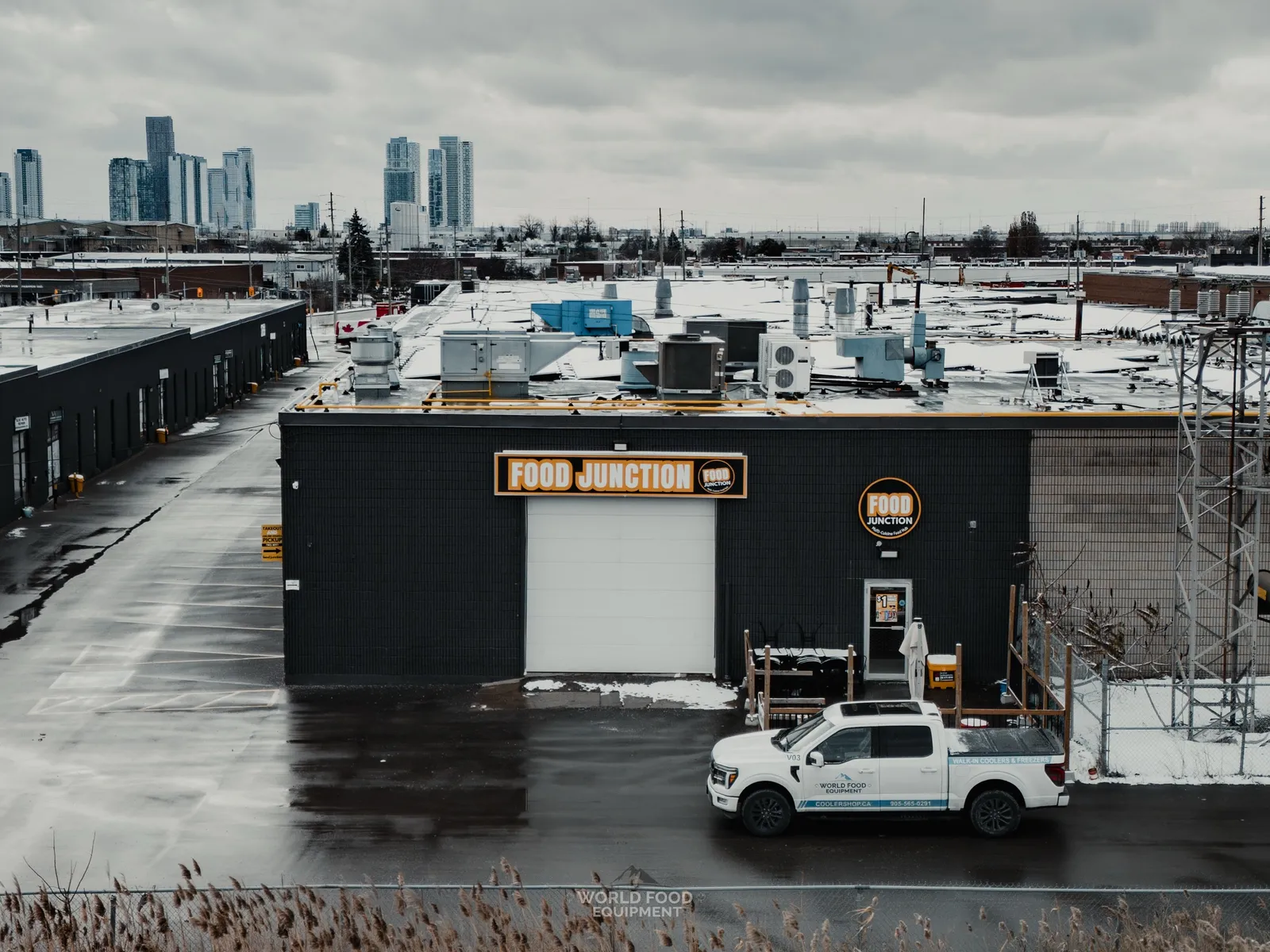 Exterior view of the Food Junction restaurant in Toronto from across the parking lot, dark facade with orange FOOD JUNCTION signage, WFE service truck parked out front, rooftop refrigeration condensers visible above and downtown Toronto skyline in the distance.