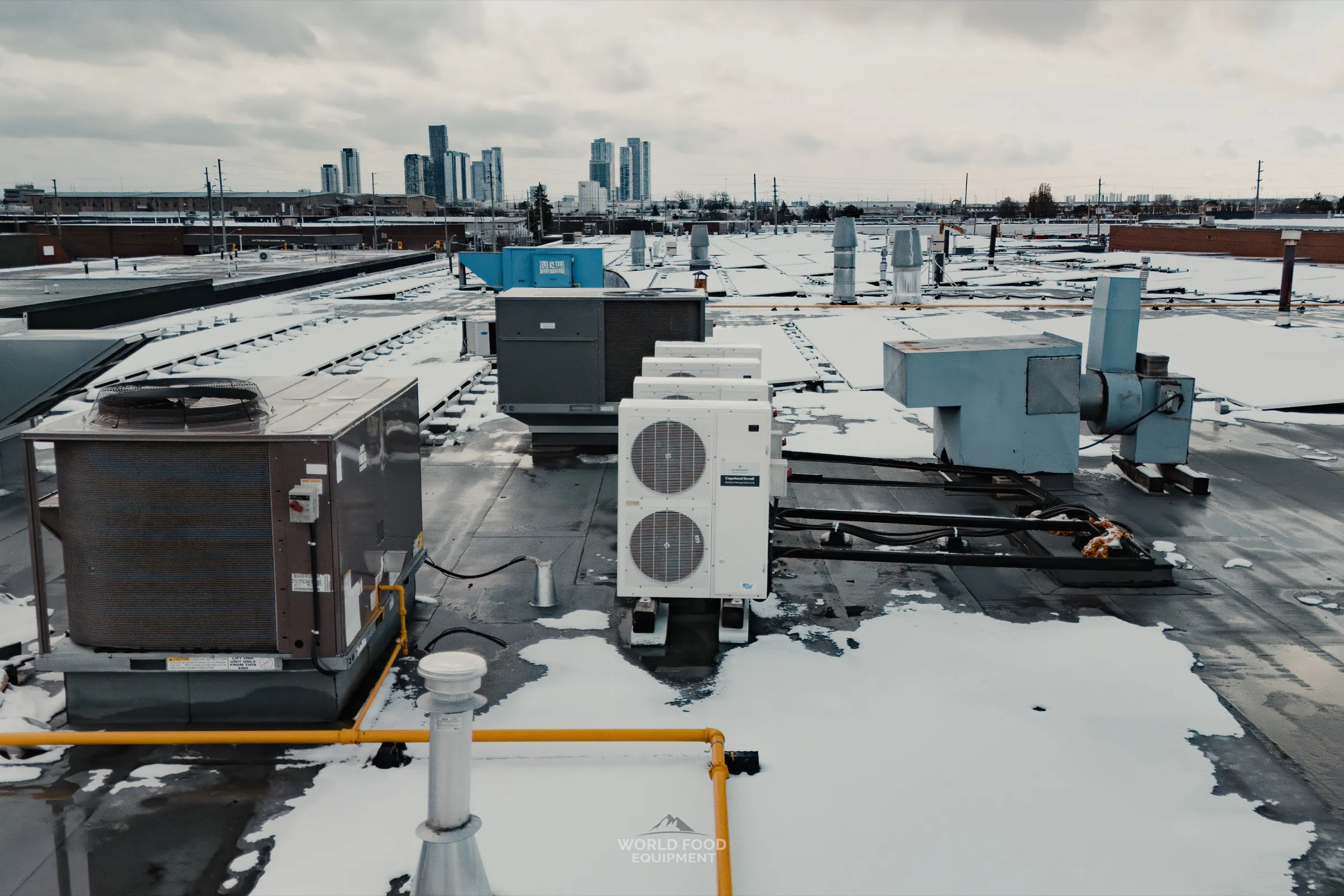 Rooftop of a Mississauga food hall after a World Food Equipment install: a Copeland Scroll outdoor refrigeration condensing unit on rubber isolators, a packaged rooftop HVAC unit, an exhaust fan, and a yellow-painted gas line across a snow-dusted flat roof