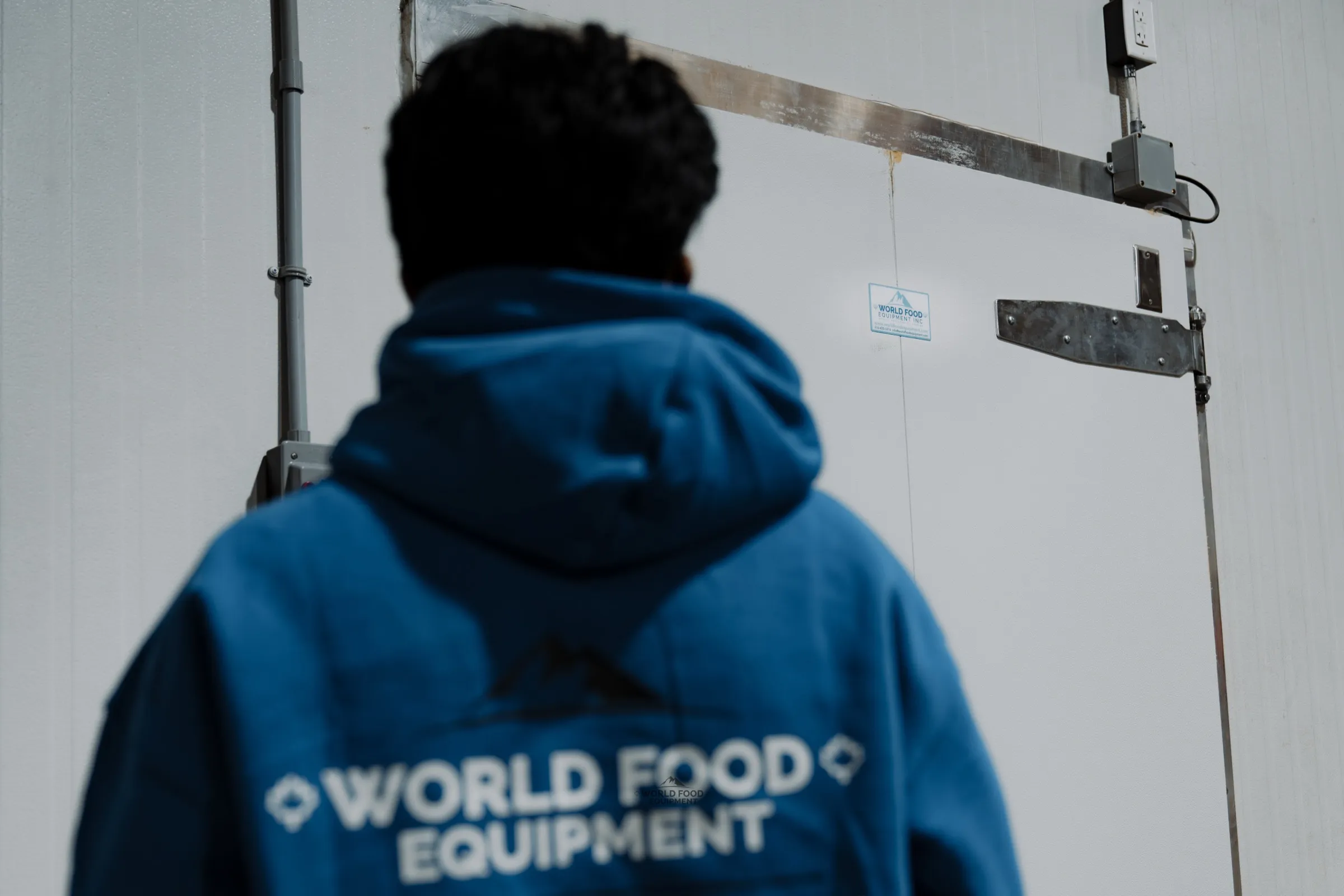 World Food Equipment crew member in a blue WFE hoodie at a walk-in cooler door at the Food Junction install in Mississauga, Ontario, with EMT conduit and a junction box mounted on the white panel wall above the door hardware