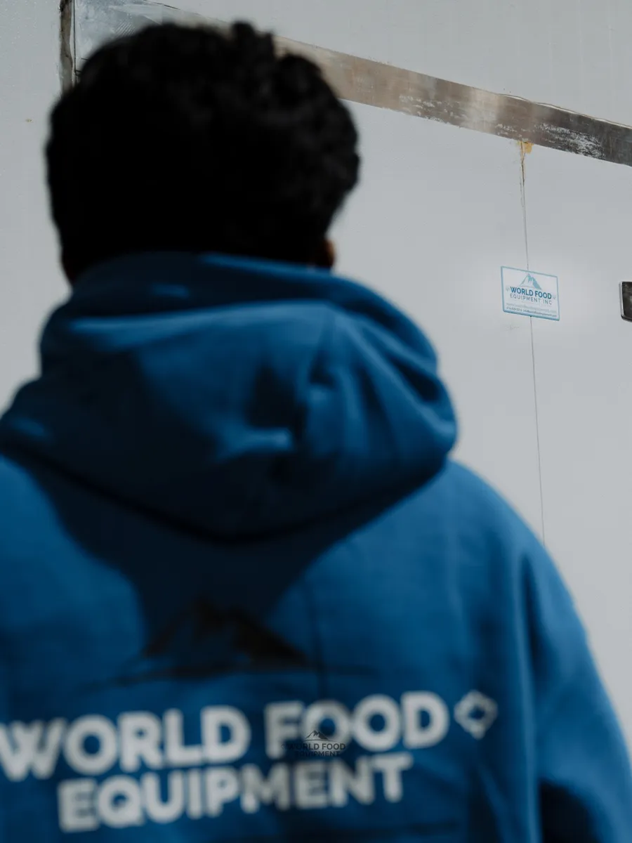 WFE installer viewed from behind in a branded blue World Food Equipment hoodie, facing a finished commercial walk-in cooler exterior with a WFE Inc decal on the panel and heavy-duty insulated door hardware, Food Junction install, Toronto, Ontario.