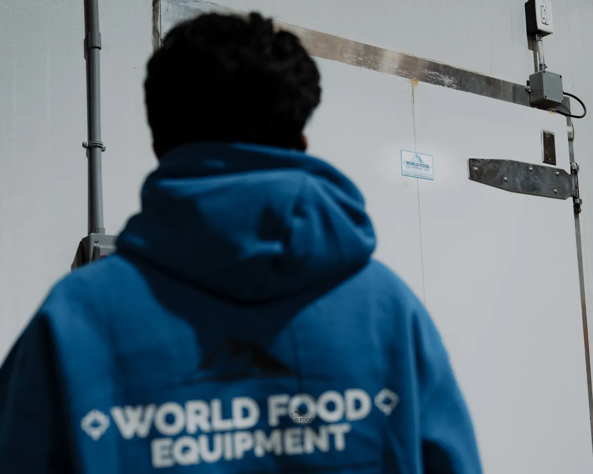 World Food Equipment installer in a blue branded hoodie viewed from behind, facing a finished white insulated cold-storage door with a stainless hinge and control box, Food Junction restaurant install, Toronto, World Food Equipment