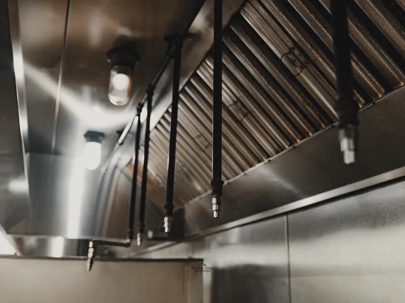 Close-up of the stainless steel kitchen exhaust hood at Food Junction Toronto with slatted filter vents and overhead light fixtures, mounted above the cooking line.