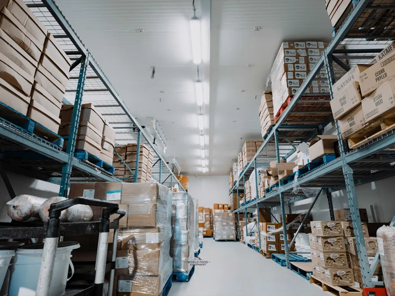 Stocked walk-in cold storage aisle at Food Junction Toronto, pendant lighting overhead, product boxes stacked on metal shelving running floor to ceiling down both sides, white panel walls, cooler operational.
