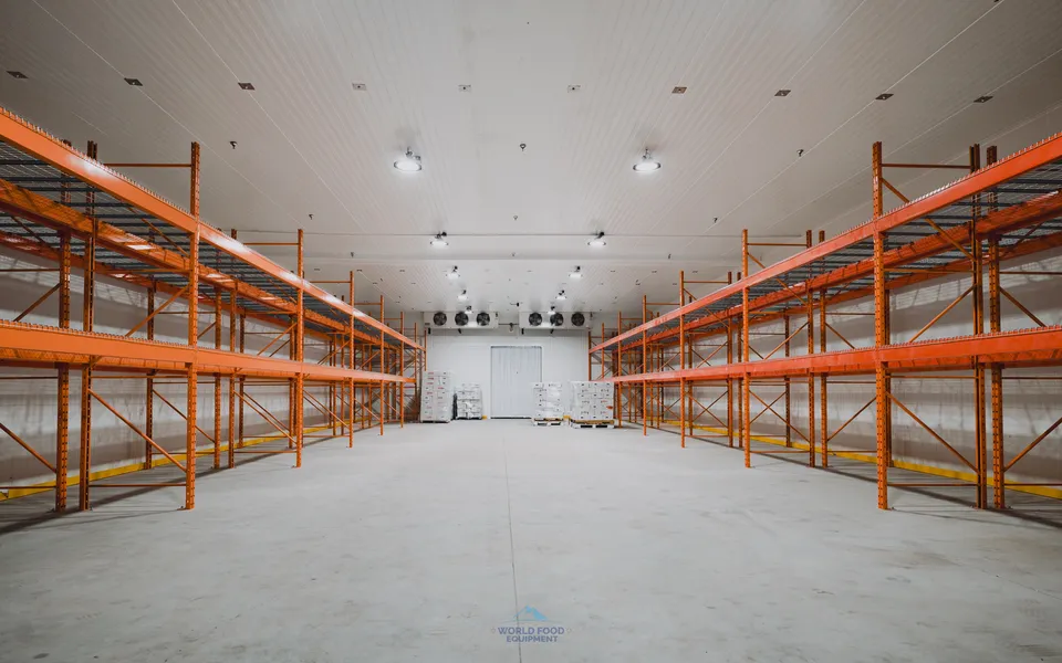 Cathedral-scale commercial cold storage facility interior with industrial orange pallet racking installed on both sides framing a central polished concrete aisle, ceiling-mounted refrigeration evaporators visible across the back wall, white insulated panel walls and ceiling, Ontario custom cold storage by World Food Equipment