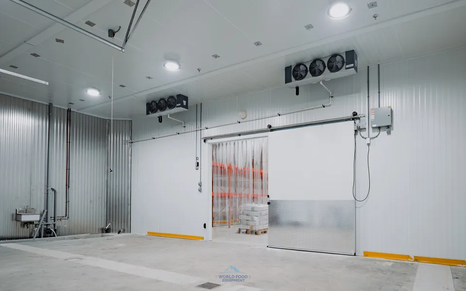 Finished commercial walk-in freezer with insulated sliding door, vinyl strip curtain to contain cold air, dual ceiling-mounted KeepRite evaporators, and yellow impact bollards at the entry, Halal Meat install, Ontario, World Food Equipment