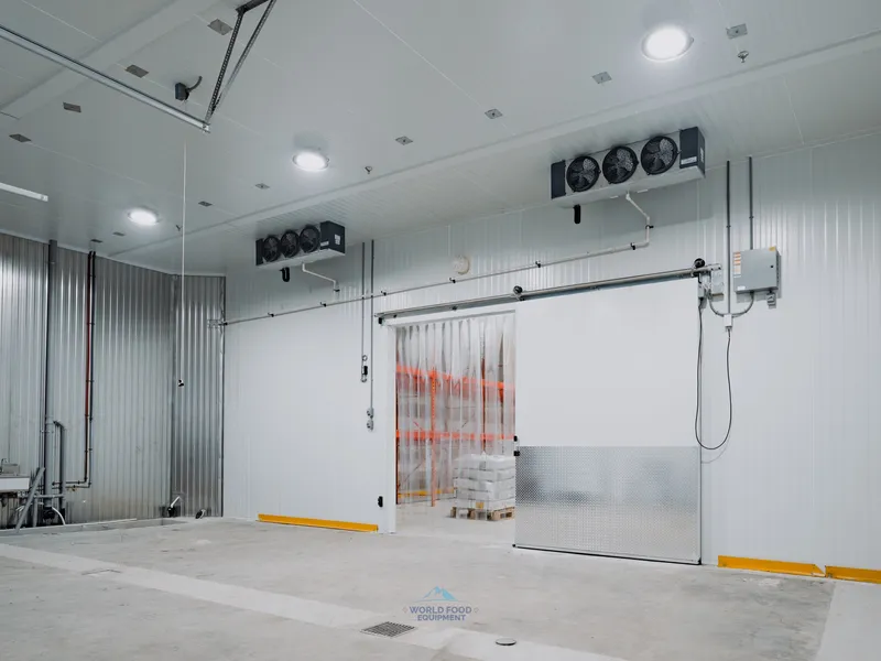 Operational walk-in freezer at the halal meat processor in Mississauga with closed sliding insulated door, vinyl strip curtain visible inside the entry, dual ceiling-mounted KeepRite evaporators above, small utility sink station to the left, polished concrete floor.