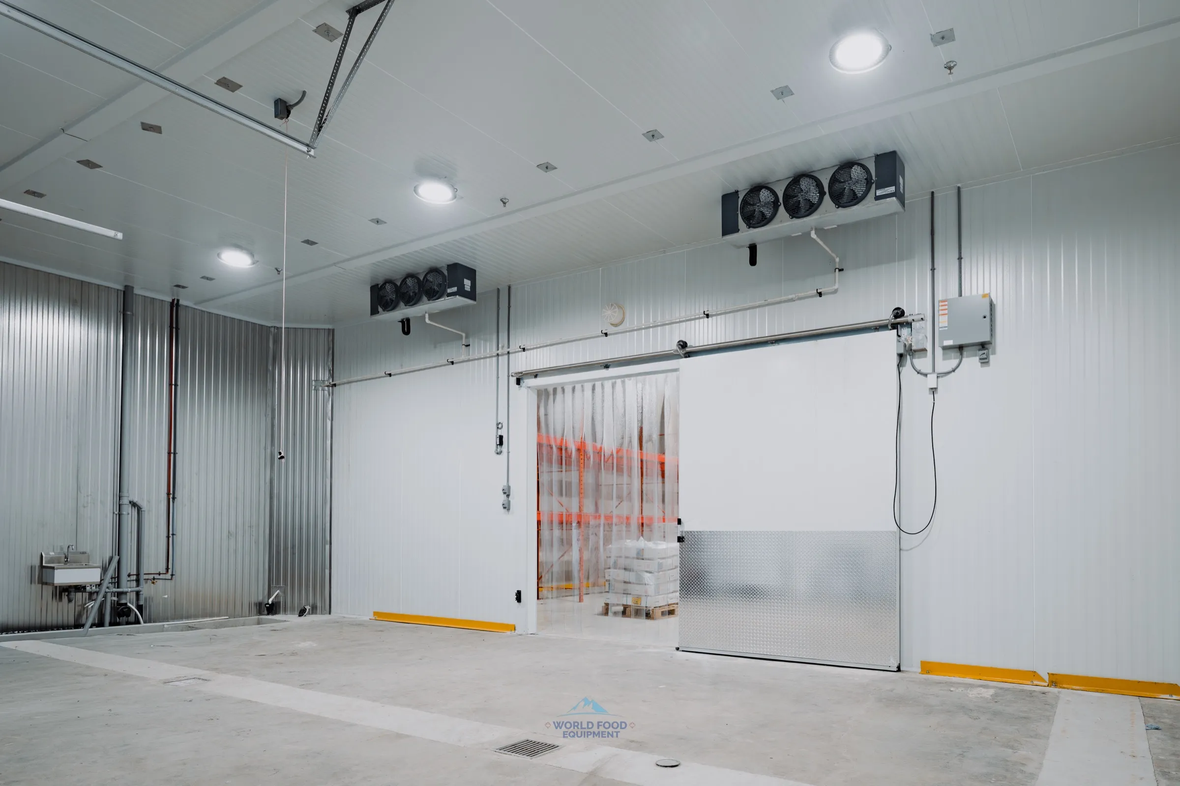 Interior of the finished halal meat processing facility cold-storage build by World Food Equipment. Insulated white sliding cooler door with orange PVC strip curtain across the opening, twin ceiling-mounted KeepRite evaporator units overhead, stainless wash-down sink and corrugated panel on the side wall, polished concrete floor