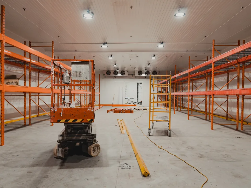 Mid-build interior of the halal meat processor cold envelope: scissor lift mid-room, orange pallet racking installed along both walls, ceiling-mounted evaporators visible at the back wall, white insulated panel walls and ceiling already in place.
