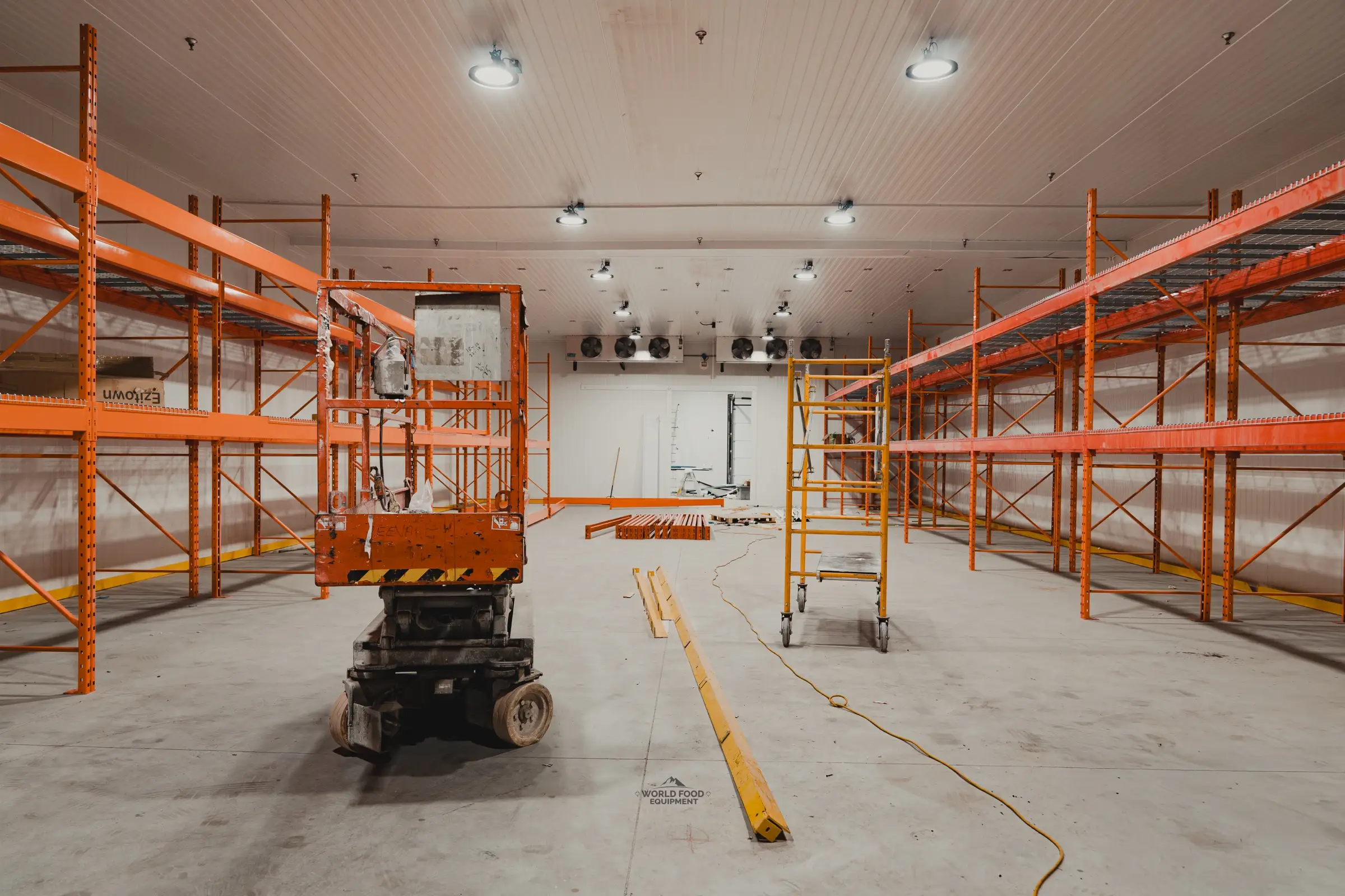 Mid-build interior of the halal meat processor cold envelope in Mississauga: scissor lift mid-room, orange pallet racking installed along both walls, ceiling-mounted evaporators visible at the back wall, white insulated panel walls and ceiling already in place, refrigeration not yet commissioned.