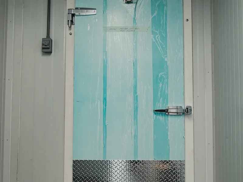 Newly installed insulated swing door at the halal meat processor with protective film still on the door panels, diamond-plate kick guard at the bottom, industrial latch hardware, white insulated panel walls flanking the entry.