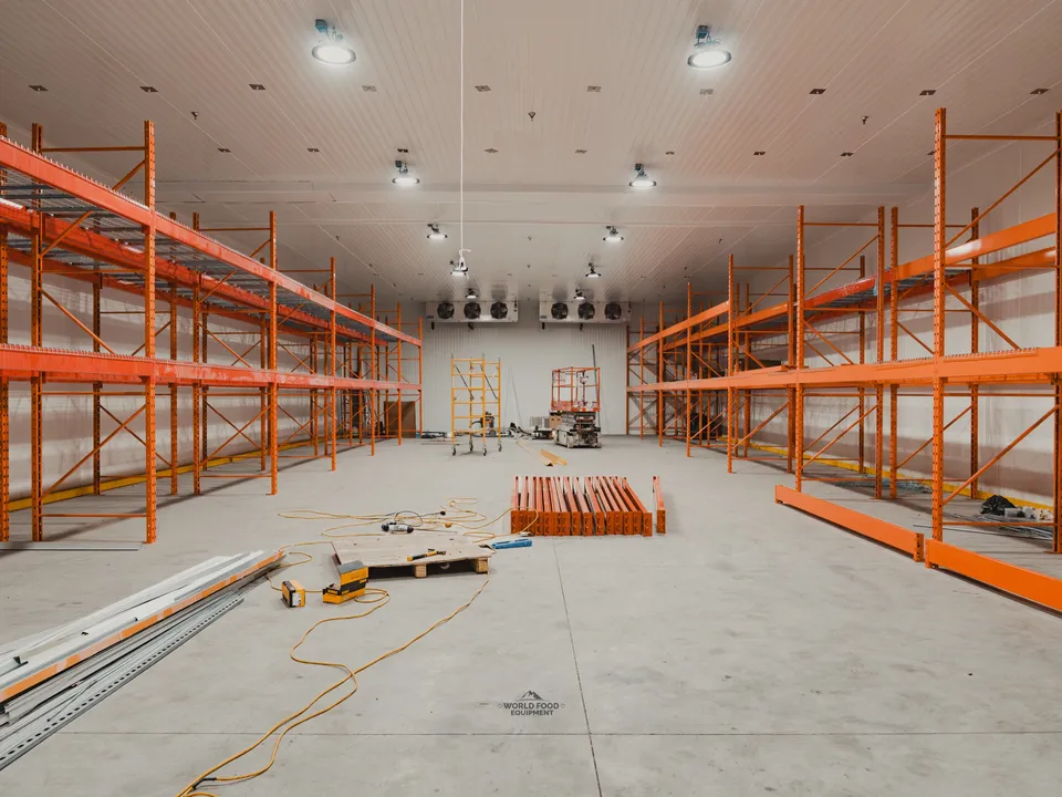 Wide interior of the halal meat processor cold envelope post-panel set: orange pallet racking installed floor to ceiling on both sides, ceiling-mounted evaporators in place at the back wall, refrigeration lines staged on the floor awaiting connection, scaffold tower mid-room.