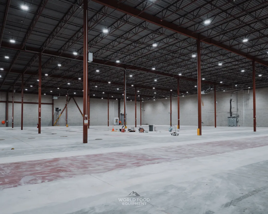 Empty industrial distribution bay with exposed red structural steel columns, polished concrete floor, and exposed steel-deck ceiling with linear LED lights after refrigeration equipment removal, Longo's decommissioning, Ontario, World Food Equipment