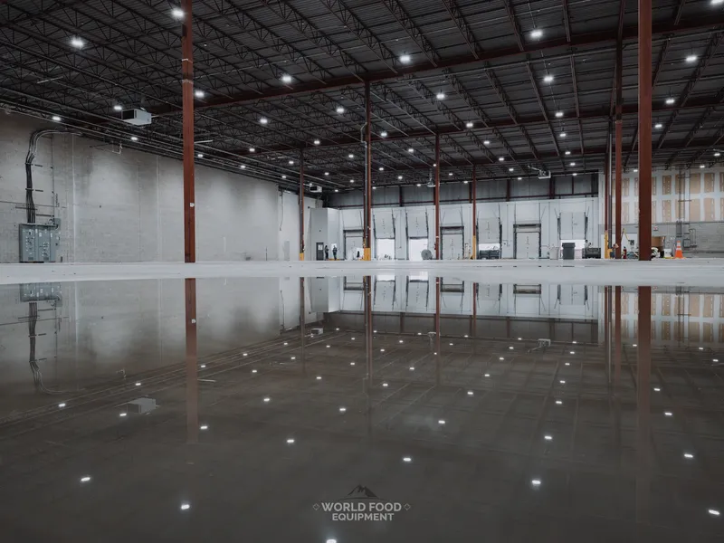 Wide view of a cleared industrial warehouse after refrigeration decommissioning, polished concrete floor reflecting the overhead lights, steel support columns down both sides, and loading dock doors at the far end, Longo's, Toronto-area, World Food Equipment