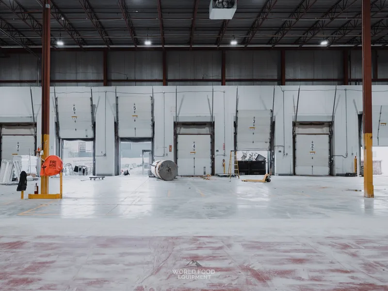 Loading dock at the Longo's Scarborough site with multiple loading bay doors in a row at the back wall, two WFE crew on the left foreground, decommissioned refrigeration units staged on the floor mid-room ready to ship out.