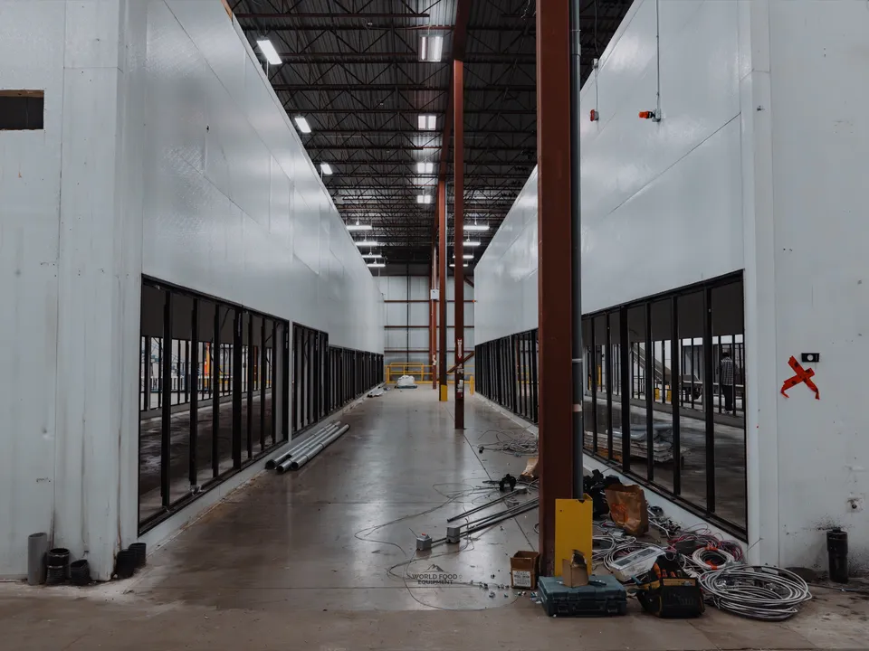 Mid-decommissioning aisle at the Longo's Scarborough distribution site: long corridor between two cooler walls with refrigerated display cases already removed, glass corridor walls still standing on both sides, debris and refrigerant lines on the floor.