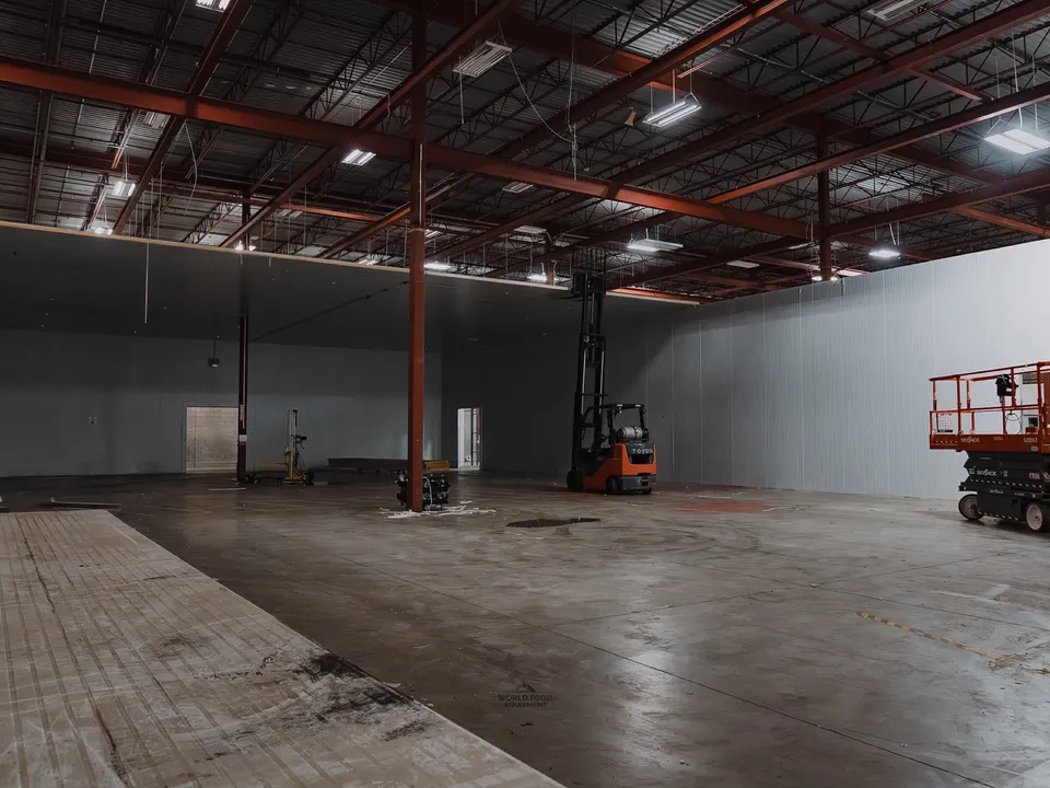 Active demolition inside the Longo's Scarborough warehouse: forklift mid-room, scissor lift staged to the right, cooler panel walls partially removed exposing the building structure behind, debris from removed panels stacked on the floor.