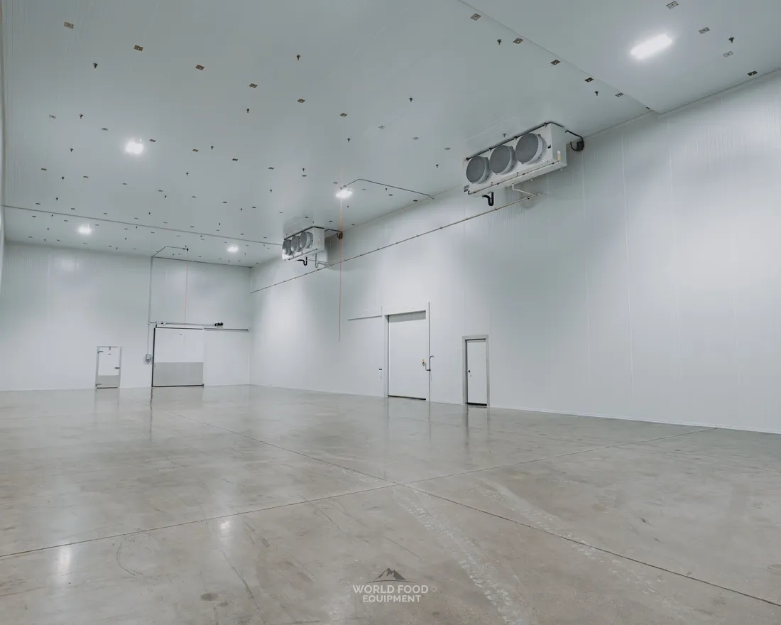 Cathedral-scale industrial cold storage envelope with finished white insulated panel walls and ceiling, two ceiling-mounted evaporators, polished concrete floor, and twin insulated walk-in doors at the back wall, Pacer Air Freight install, Mississauga, World Food Equipment